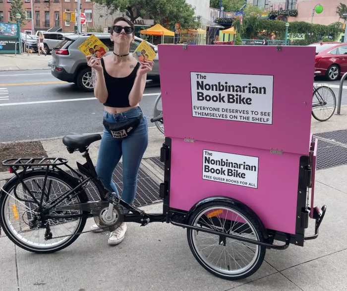 person with black cargo bike with pink cargo boxes labeled "nonbinarian book bike"